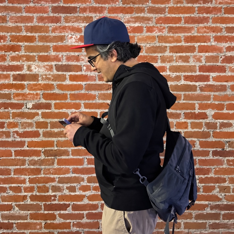 Karna, in a navy cap and black hoodie, looking at a phone in front of a brick wall.